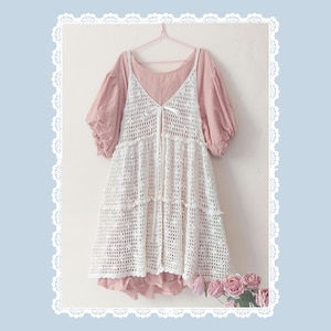 Puede incluir: Un conjunto de capas que presenta una parte superior rosa claro con mangas cortas abullonadas, combinada con un vestido de ganchillo blanco. El vestido de ganchillo tiene cuello en V y falda escalonada. Un pequeño lazo blanco adorna el frente. El conjunto está colgado de una percha rosa.