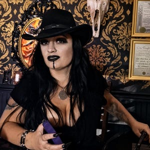 Pode incluir: Um chapéu fedora preto com uma faixa fina, usado por uma pessoa com cabelos longos e escuros, batom escuro e uma linha vertical no queixo. A pessoa veste uma blusa preta e um colar com pingente redondo, segurando um objeto roxo.