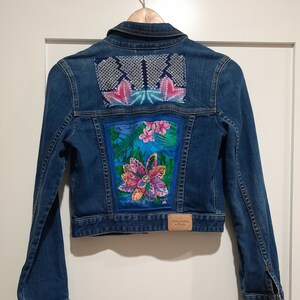 May include: A blue denim jacket with a floral and geometric design on the back. The jacket has a label that says "Abercrombie & Fitch" on the bottom right side of the back.