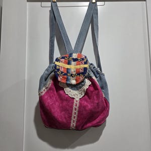 May include: A backpack constructed from light blue denim and vibrant magenta fabric. The bag features a flap with a multi-colored plaid pattern, a white button closure, and decorative white lace trim accents along the front panel and straps.