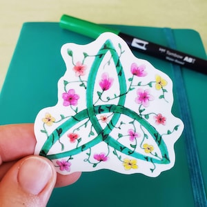 May include: A green Celtic knot design on a white background. The knot is surrounded by vines with pink, yellow and white flowers.