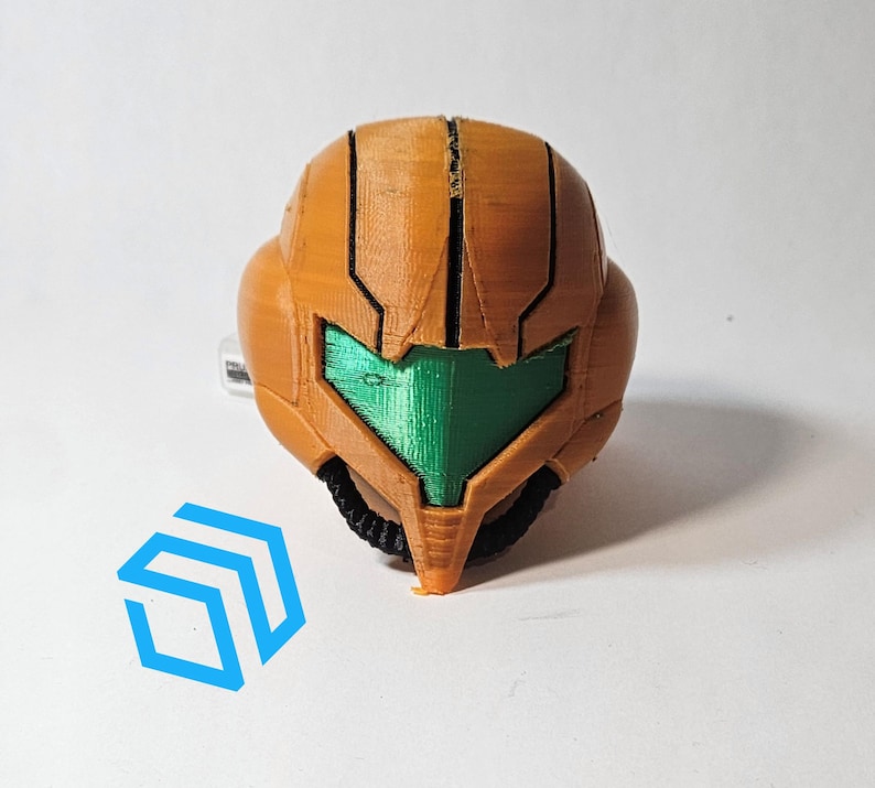 Pode incluir: Um capacete laranja e verde impresso em 3D com uma viseira. O capacete &eacute; uma r&eacute;plica de um personagem de um videojogo.