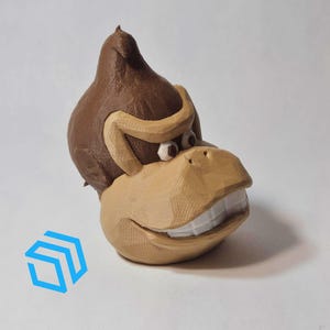 Pode incluir: Uma figurinha de um macaco marrom impressa em 3D com um largo sorriso. O macaco usa um gorro de pele marrom e tem dentes grandes e brancos.