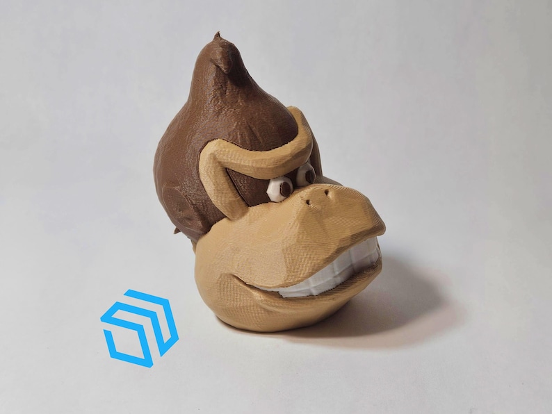Pode incluir: Uma figurinha de um macaco marrom impressa em 3D com um largo sorriso. O macaco usa um gorro de pele marrom e tem dentes grandes e brancos.