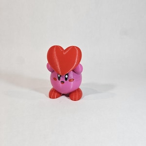 Pode incluir: Uma figura de um personagem de desenho animado impressa em 3D em rosa e vermelho, segurando um cora&ccedil;&atilde;o vermelho. O personagem tem um corpo redondo, bra&ccedil;os e pernas pequenos e uma cabe&ccedil;a grande com uma express&atilde;o de surpresa.