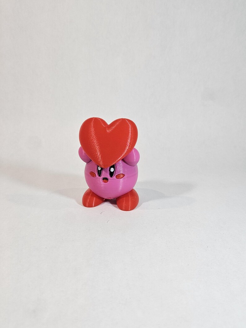Pode incluir: Uma figura de um personagem de desenho animado impressa em 3D em rosa e vermelho, segurando um cora&ccedil;&atilde;o vermelho. O personagem tem um corpo redondo, bra&ccedil;os e pernas pequenos e uma cabe&ccedil;a grande com uma express&atilde;o de surpresa.