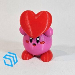 Pode incluir: Uma figura impressa em 3D rosa e vermelha de um personagem segurando um cora&ccedil;&atilde;o. O personagem tem um corpo redondo, bra&ccedil;os e pernas pequenos e um rosto bonito.