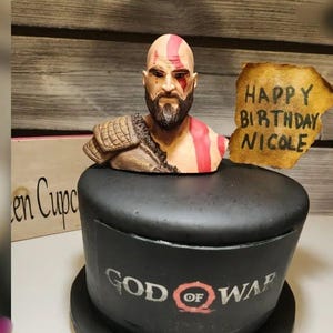 May include: A black cake decorated with a fondant figure of Kratos from the God of War video game. The cake is topped with a brown paper sign that says "Happy Birthday Nicole".