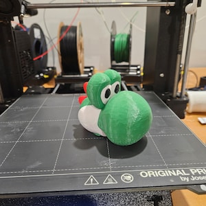 Pode incluir: Uma figura de Yoshi, um personagem da s&eacute;rie de videogames Super Mario, impressa em 3D em verde e branco. A figura est&aacute; sentada em uma cama de impressora preta com uma tela digital que mostra o progresso da impress&atilde;o.
