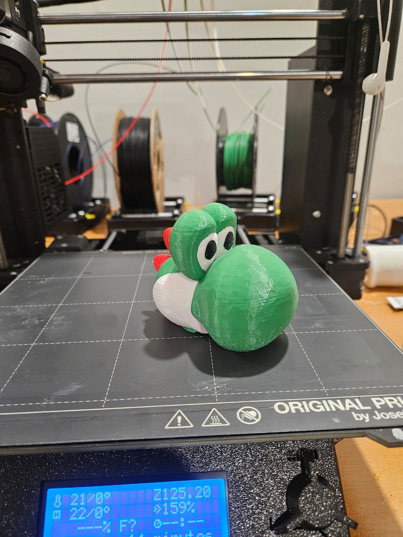 Pode incluir: Uma figura de Yoshi, um personagem da s&eacute;rie de videogames Super Mario, impressa em 3D em verde e branco. A figura est&aacute; sentada em uma cama de impressora preta com uma tela digital que mostra o progresso da impress&atilde;o.