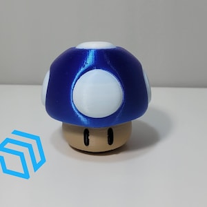 Könnte beinhalten: Eine blaue und weiße Pilzfigur mit braunem Sockel. Der Pilz hat weiße Punkte auf der Oberseite und zwei schwarze Augen auf dem Sockel.