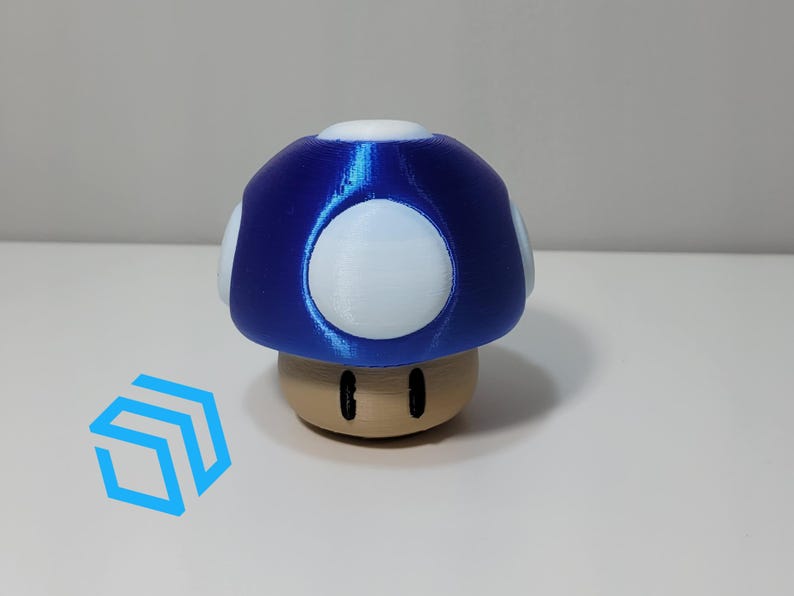 Pode incluir: Uma figurinha em forma de cogumelo azul e branco com uma base marrom. O cogumelo tem manchas brancas no topo e dois olhos pretos na base.