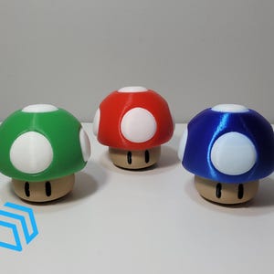 Pode incluir: Tr&ecirc;s figurinhas de cogumelo impressas em 3D, uma vermelha, uma verde e uma azul. Cada cogumelo tem manchas brancas e um caule castanho.