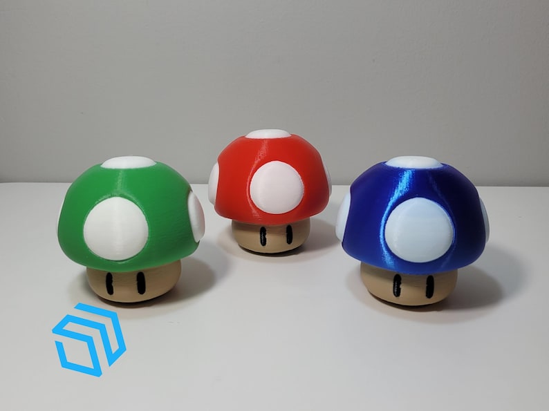 Pode incluir: Tr&ecirc;s figurinhas de cogumelo impressas em 3D, uma vermelha, uma verde e uma azul. Cada cogumelo tem manchas brancas e um caule castanho.