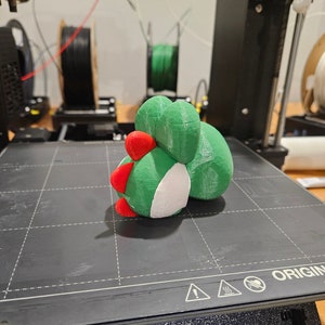 Pode incluir: Um objeto impresso em 3D de cor verde, branca e vermelha sobre uma superf&iacute;cie preta. O objeto tem a forma de um personagem de desenho animado com um corpo redondo e uma cabe&ccedil;a pequena. O objeto est&aacute; sentado sobre uma superf&iacute;cie preta com um padr&atilde;o de grade.