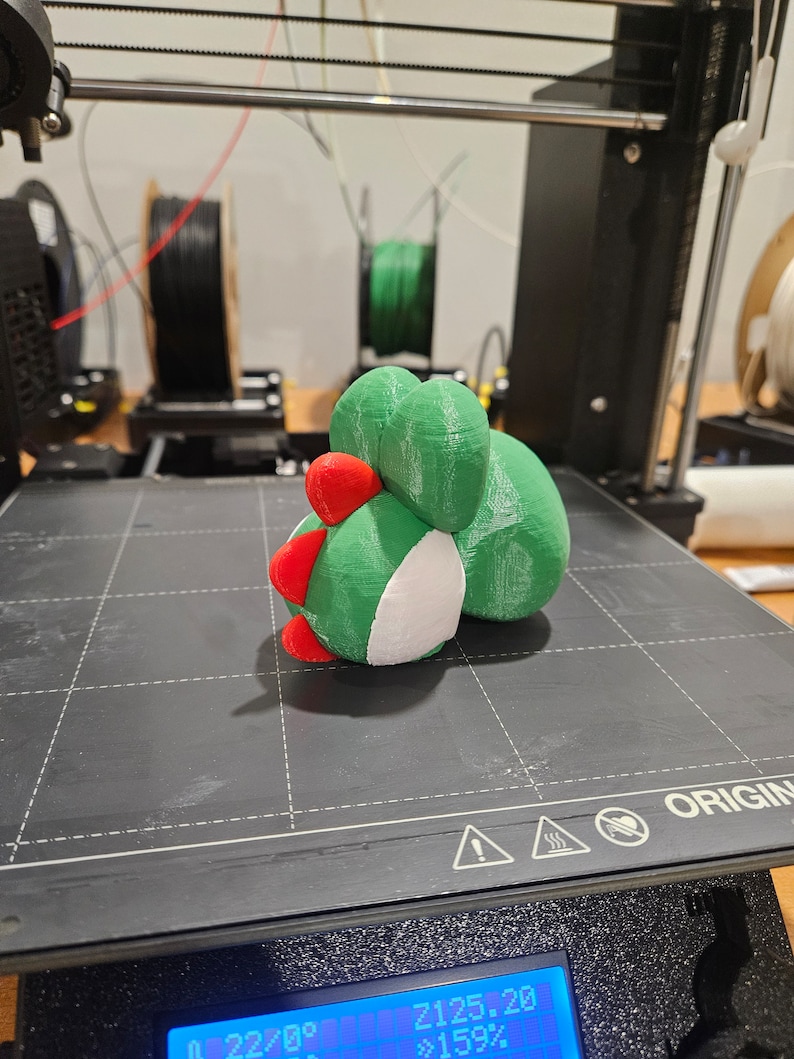 Pode incluir: Um objeto impresso em 3D de cor verde, branca e vermelha sobre uma superf&iacute;cie preta. O objeto tem a forma de um personagem de desenho animado com um corpo redondo e uma cabe&ccedil;a pequena. O objeto est&aacute; sentado sobre uma superf&iacute;cie preta com um padr&atilde;o de grade.