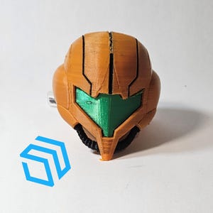 Pode incluir: Um capacete impresso em 3D de cor laranja e verde com uma viseira. O capacete &eacute; uma r&eacute;plica de um personagem de um videojogo.
