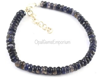 May include: A blue gemstone bracelet with a silver clasp. The bracelet is made of small, faceted beads. OpalGemsEmporium is printed on the white background.