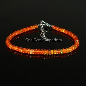Könnte beinhalten: Ein zartes Armband aus orangefarbenen Opalperlen mit einem silbernen Verschluss. Das Armband besteht aus kleinen, runden Perlen, die auf einer dünnen silbernen Kette aufgereiht sind. Die Perlen haben eine leuchtende orange Farbe mit einem leichten irisierenden Schimmer. Der Verschluss ist ein einfacher, silberner Haken- und Ösenverschluss.