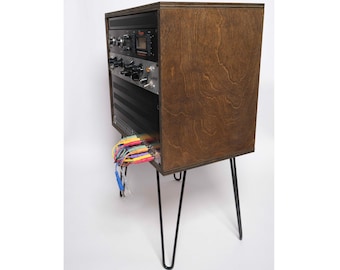 May include: A wooden rack with black metal legs, holding a rack of audio equipment. The rack has a brown wood finish and the legs are hairpin style. A bundle of colorful cables is hanging from the bottom of the rack.