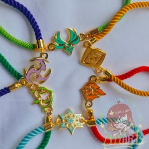 Op de afbeelding: Zeven kleurrijke armbandjes van touw met gouden bedels in verschillende ontwerpen. De bedels zijn in de vorm van een ster, een hart, een vlam, een werveling, een vierkant, een vogel en een sneeuwvlok.