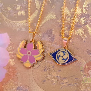 Puede incluir: Dos collares de oro con colgantes. Un colgante es una flor rosa con detalles amarillos. El otro colgante es un ojo azul con remolinos dorados.