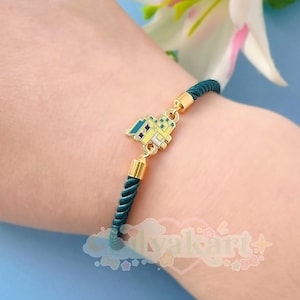 Puede incluir: Un brazalete de cuerda verde con un dije dorado de una casa amarilla con detalles azules.