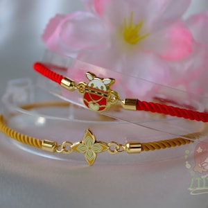 Puede incluir: Dos pulseras con detalles dorados. Una es roja con un dije dorado con un animal de dibujos animados en una bolsa roja. La otra es dorada con un dije de trébol de cuatro hojas. Ambas están sobre un expositor transparente, con una flor rosa de fondo.