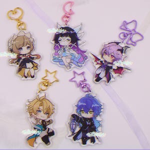 Puede incluir: Cinco llaveros de personajes de estilo anime con herrajes dorados o rosas. Los personajes tienen varios colores de pelo y atuendos, incluyendo una sirvienta, un hada, un vampiro y dos figuras masculinas. Cada llavero es un diseño diferente.