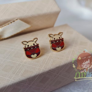 Puede incluir: Un par de pendientes de botón de tono dorado con un diseño de animal de dibujos animados. El animal lleva un suéter rojo y blanco con un corazón en la parte delantera. Los pendientes están en una pequeña caja de regalo.