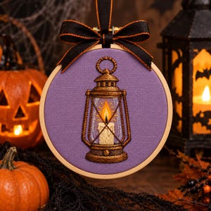 Peut inclure: Un tambour à broder circulaire en bois contenant un panneau en tissu violet avec un motif brodé à la main représentant une lanterne brune avec une bougie allumée à la flamme jaune et orange. Un ruban noir à bordure orange est noué en haut.