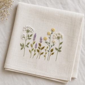 May include: A square off-white fabric napkin featuring detailed embroidery of various wildflowers. The design includes white umbel flowers, purple lavender-like stalks, and small yellow blossoms with green stems and leaves centered on the cloth.