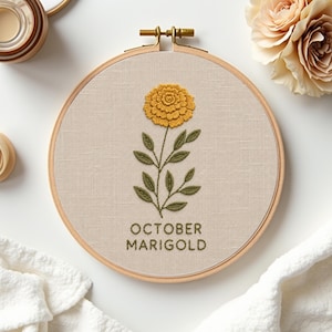 May include: A circular wooden embroidery hoop containing beige fabric with a hand-stitched yellow marigold flower, green leaves, and the text OCTOBER MARIGOLD embroidered in green thread centered below the floral design on the fabric.