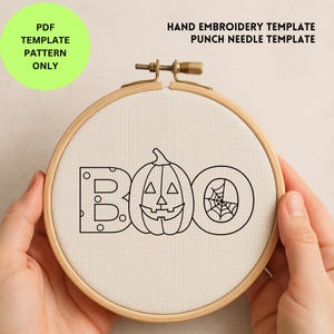 May include: A wooden embroidery hoop with a canvas featuring a black outline design of the word "BOO" with a jack-o'-lantern and spiderweb. Text on the left says "PDF TEMPLATE PATTERN ONLY". The text at the top says "HAND EMBROIDERY TEMPLATE PUNCH NEEDLE TEMPLATE".
