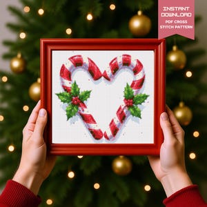 May include: A framed cross-stitch pattern featuring a heart made of red and white candy canes, adorned with green holly and red berries. The frame is red, and the text "INSTANT DOWNLOAD PDF CROSS STITCH PATTERN" is visible.
