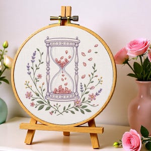May include: A circular wooden embroidery hoop containing a white fabric panel with a cross-stitched hourglass design. The hourglass is filled with pink petals, surrounded by embroidered lavender sprigs, green leaves, and small pink flowers.