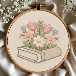 Può includere: Un telaio da ricamo circolare in legno contenente un tessuto color bianco sporco con un disegno ricamato a mano di un libro chiuso. Un bouquet di tulipani rosa, margherite rosa chiaro, piccoli fiori blu e foglie verdi emerge dal libro.