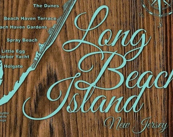 May include: A wooden sign with a map of Long Beach Island, New Jersey. The map is outlined in teal blue and includes the names of various towns and beaches on the island. The words "Long Beach Island" are written in a large, teal blue cursive font. The sign also includes a compass rose.