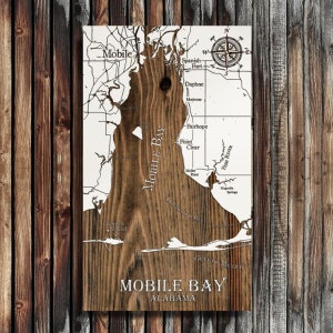 May include: Wooden wall art featuring a detailed map of Mobile Bay, Alabama. The map is carved into a dark wood grain, with white detailing for the land and water features. Includes text that reads "MOBILE BAY ALABAMA" and a compass rose.