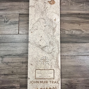 May include: A wooden map of the John Muir Trail in California, USA. The map shows the trail's length, location, trailheads, elevation change, and summit of Mount Whitney. The map is surrounded by a wooden frame.