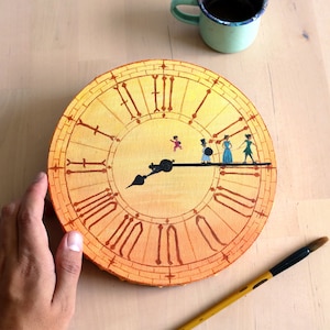 Puede incluir: Una esfera de reloj redonda pintada a mano con números romanos y un degradado amarillo y naranja. Las manecillas del reloj son negras y hay pequeñas figuras pintadas en el minutero. Un pincel y una taza de café están cerca.