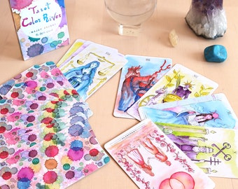 May include: A Tarot deck named "Tarot Color River" with watercolour illustrations is shown, with several cards spread out. The deck box and card backs have colourful ink blot designs. A crystal ball, amethyst cluster, and other crystals are also present.