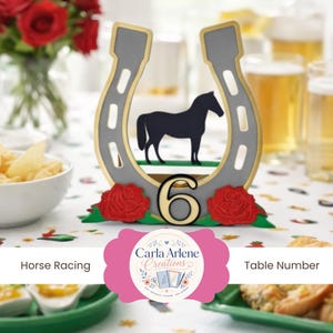May include: A table number six, featuring a grey horseshoe design with a gold trim, a black horse silhouette, and red roses. The table number is for horse racing events.
