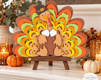May include: A layered wooden turkey cutout featuring a brown body, white collar, and a multi-colored fan tail in shades of orange, yellow, and light green. The turkey wears a black hat with a yellow buckle and stands on a small wooden easel.