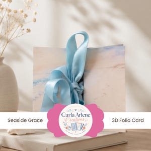Puede incluir: Una tarjeta tipo folio 3D titulada Seaside Grace de Carla Arlene Creations, que presenta una escena de playa impresa en tonos azules suaves y arena, atada con una cinta de satén azul claro. La tarjeta se muestra erguida, mostrando la imagen costera.