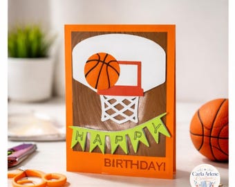 May include: A rectangular orange greeting card featuring a basketball hoop and a basketball. A green banner across the bottom displays the word HAPPY, and the word BIRTHDAY is printed in orange below the banner on the card surface.
