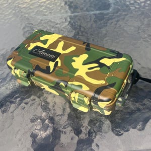 以下が含まれることがあります： カモフラージュ柄のハードケース。黒いラベルには、筆記体で「Amani」と書いてあります。