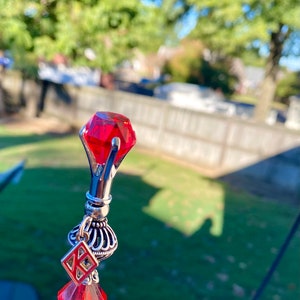 Op de afbeelding: Een zilveren haarstok met een rode kristal aan de bovenkant, een zilveren bedel met een geometrisch ontwerp en een rode kristal aan de onderkant.