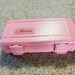 以下が含まれることがあります： ピンクのハードケースで、黒いストラップが付いています。上部に「Amani Fine Cigar Accessories」の文字があります。