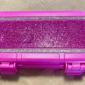 以下が含まれることがあります： クリアなラインストーンのデザインが施されたピンクの硬質プラスチックケース。ケースは前面に2つのラッチと黒いストラップが付いています。